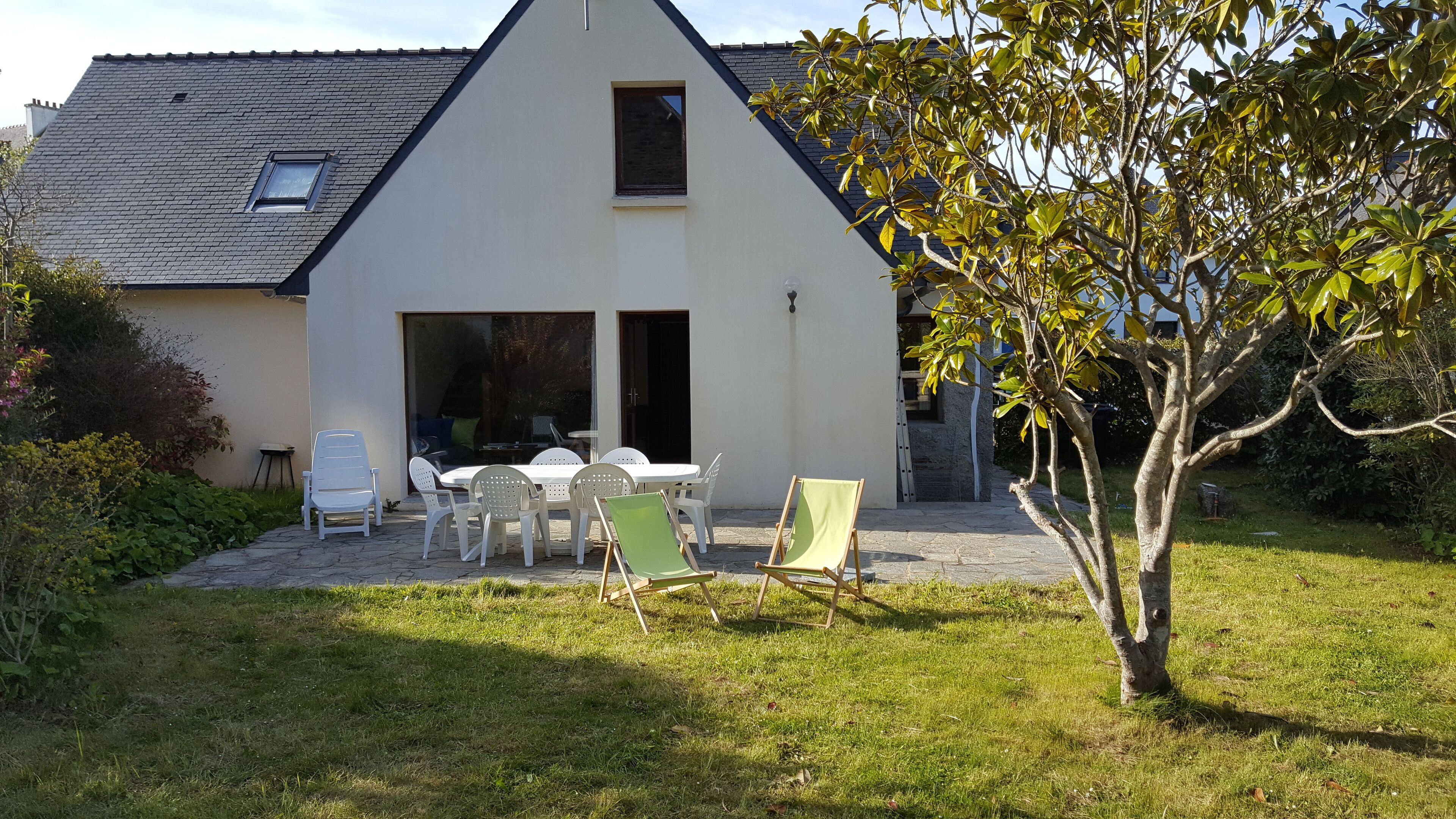 Une maison avec un toit en pente, une terrasse avec des meubles d’extérieur et un arbre au premier plan.