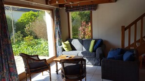 A room with a sofa, two wicker chairs, a small table, and a view of greenery outside. - Joli Cottage en Pierres Debouts Dans un Beau Jardin Fleuri Proche Plage (Nevez)