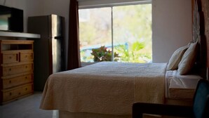 Quarto com cama, cômoda, geladeira e janela com vista para o verde.