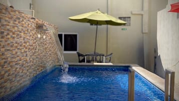 Une piscine agrémentée d’un élément décoratif aquatique, un mur recouvert de carreaux, une table avec des chaises et un parasol jaune.