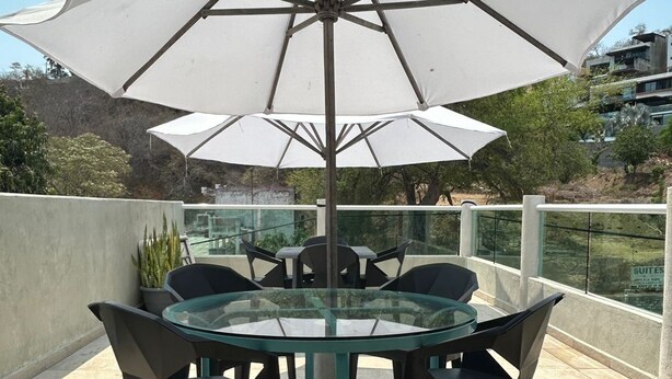 Mesa de vidro redonda com quatro cadeiras e um grande guarda-sol branco em um terraço.