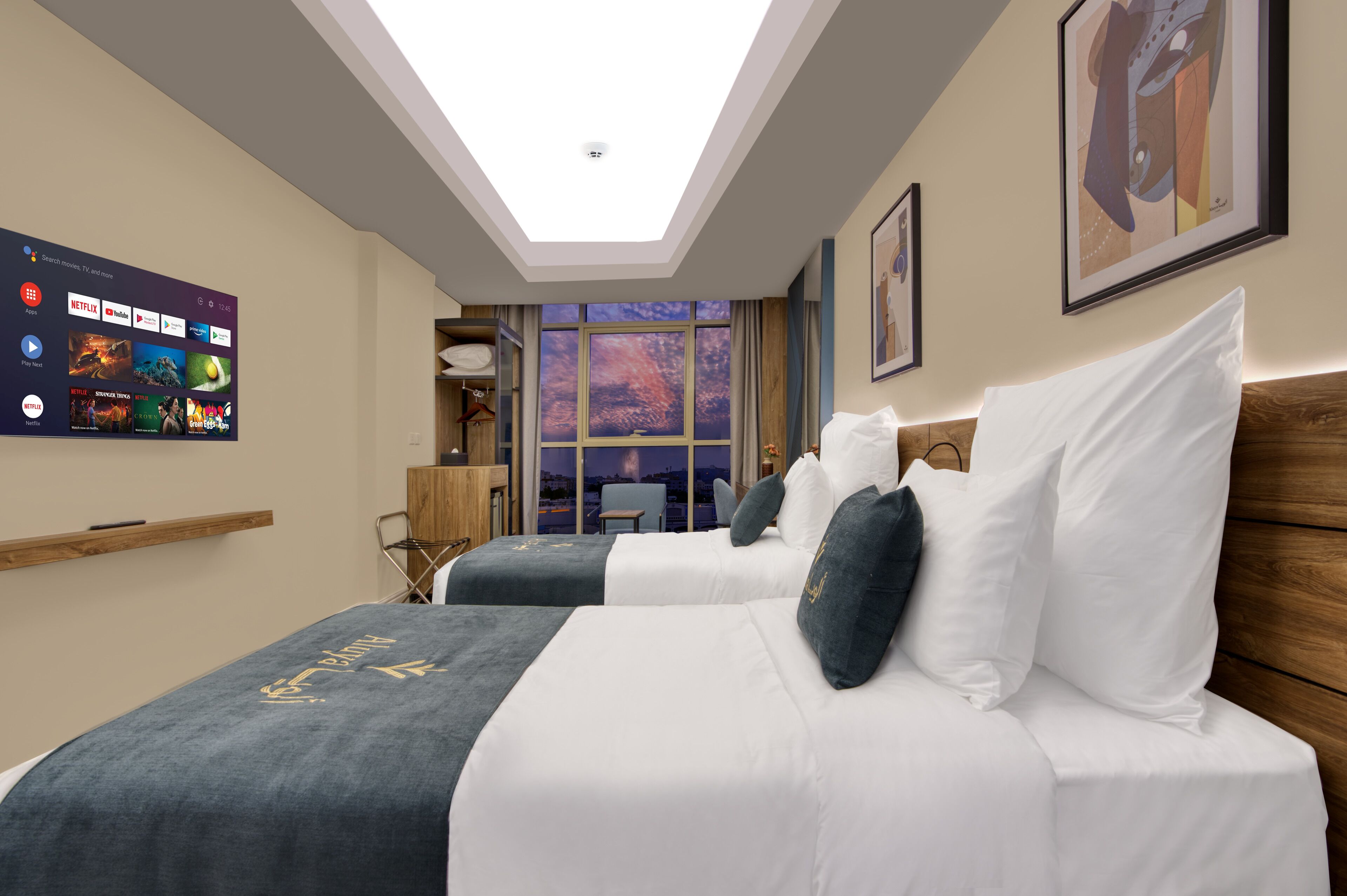 A hotel room with a skylight, a large window with a city view, a bed with white and dark bedding, and a wall-mounted TV displaying streaming services.