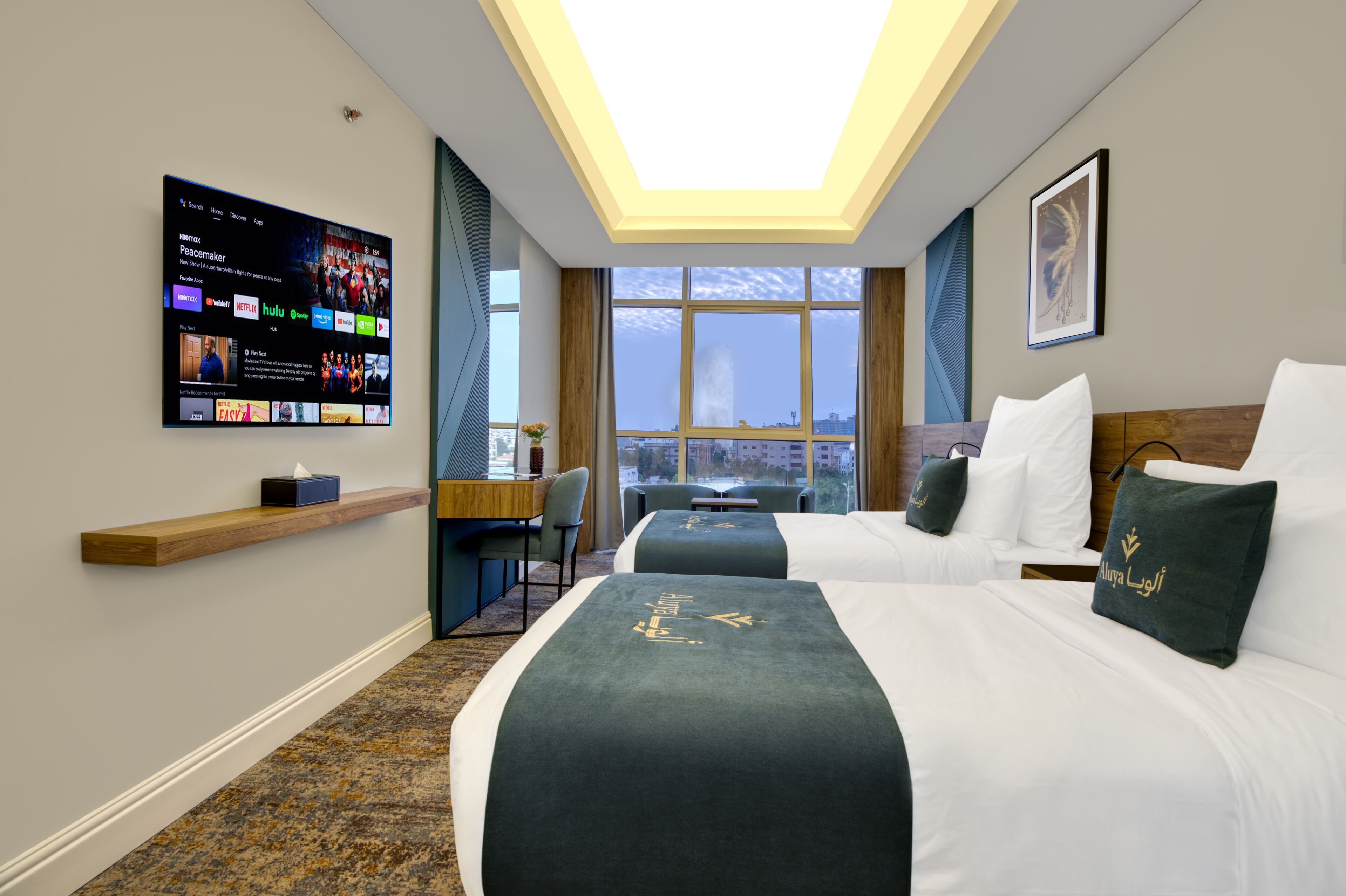 Camera d'albergo con due letti, una scrivania, una TV che mostra servizi di streaming e un'ampia finestra con vista sulla città.