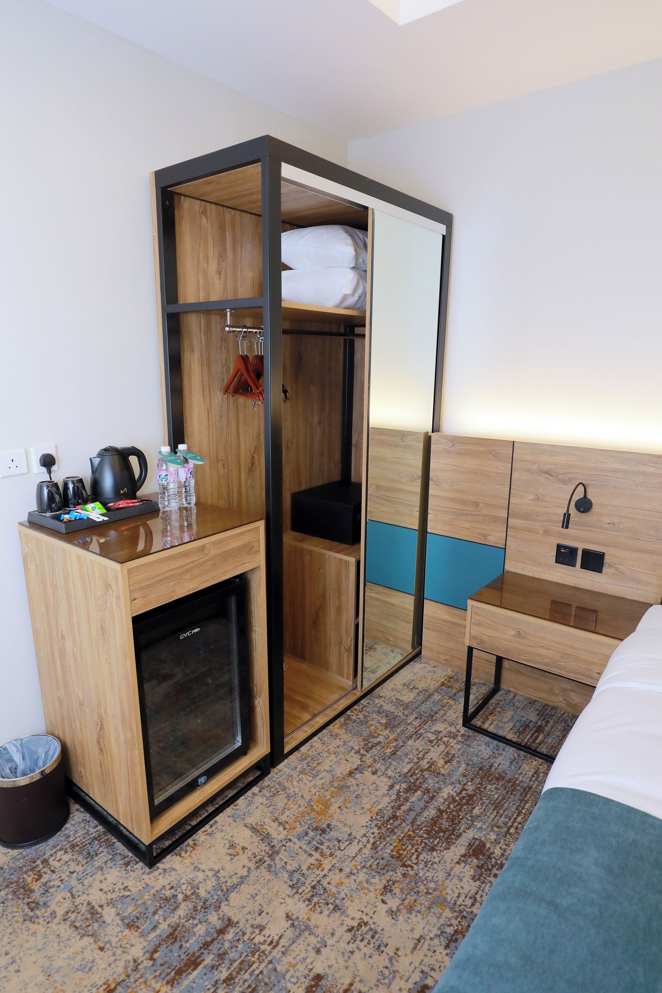 A compact hotel room with a built-in wardrobe, a bedside table with a kettle and bottles, a small fridge, and a bed with a headboard.