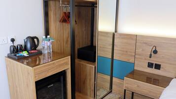 A compact hotel room with a built-in wardrobe, a bedside table with a kettle and bottles, a small fridge, and a bed with a headboard.