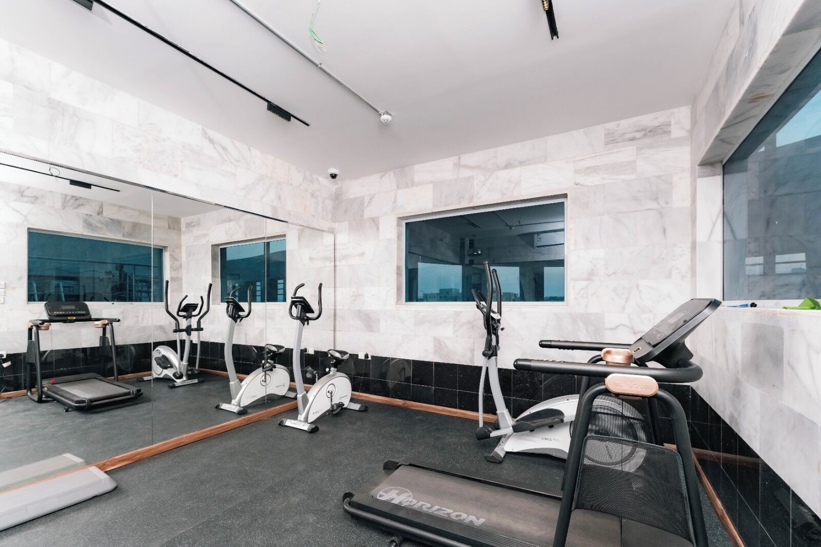 A modern gym with a variety of cardio equipment, large windows, and a marble-tiled floor.