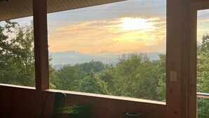 A room with a large window offering a view of a sunset over a mountainous landscape. Inside, there is a patterned rug, a green chair, and a small round table.