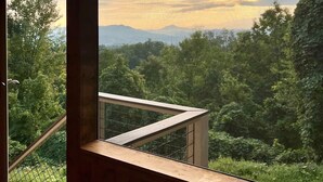A wooden balcony with a view of a forested landscape and mountains in the distance.