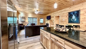 A wooden interior with a kitchen area featuring a refrigerator, a dining table with chairs, and a large window with blinds.
