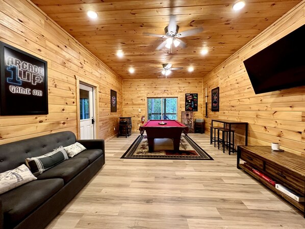 A cozy room with wooden walls, a pool table, a black sofa, and a television mounted on the wall.