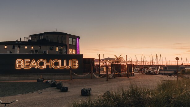A beach club with a neon sign, a building with multiple windows, and a dock area with boats. - RBR 1747 - Beach Resort Kamperland (Kamperland)