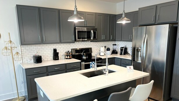 Una cocina moderna con armarios grises, electrodomésticos de acero inoxidable, una isla central con taburetes y luminarias colgantes.