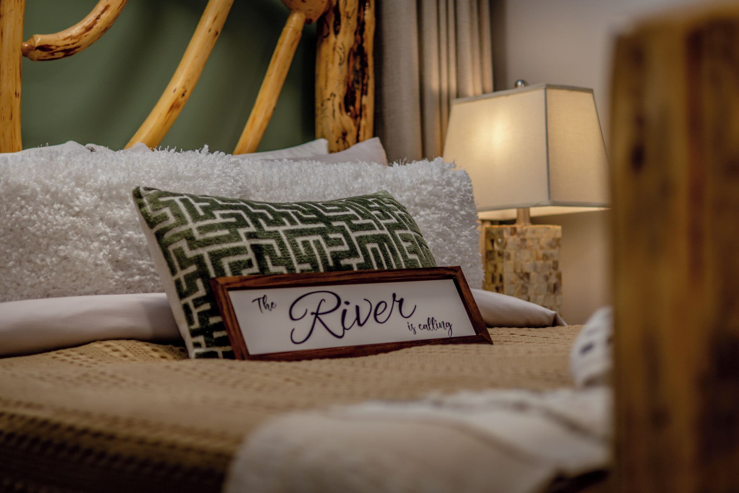 A neatly made bed with a pillow, a sign reading "The River is calling," and a lamp in the background.
