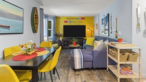 Una sala moderna con un sofá, una mesa de comedor y sillas. Hay un televisor, una estantería y un ventanal con cortinas.