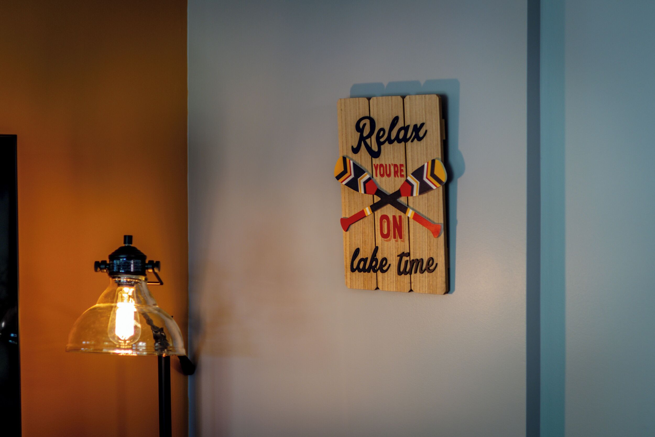 A wooden sign with the text "Relax you're on lake time" and two crossed paddles, mounted on a wall next to a lamp.
