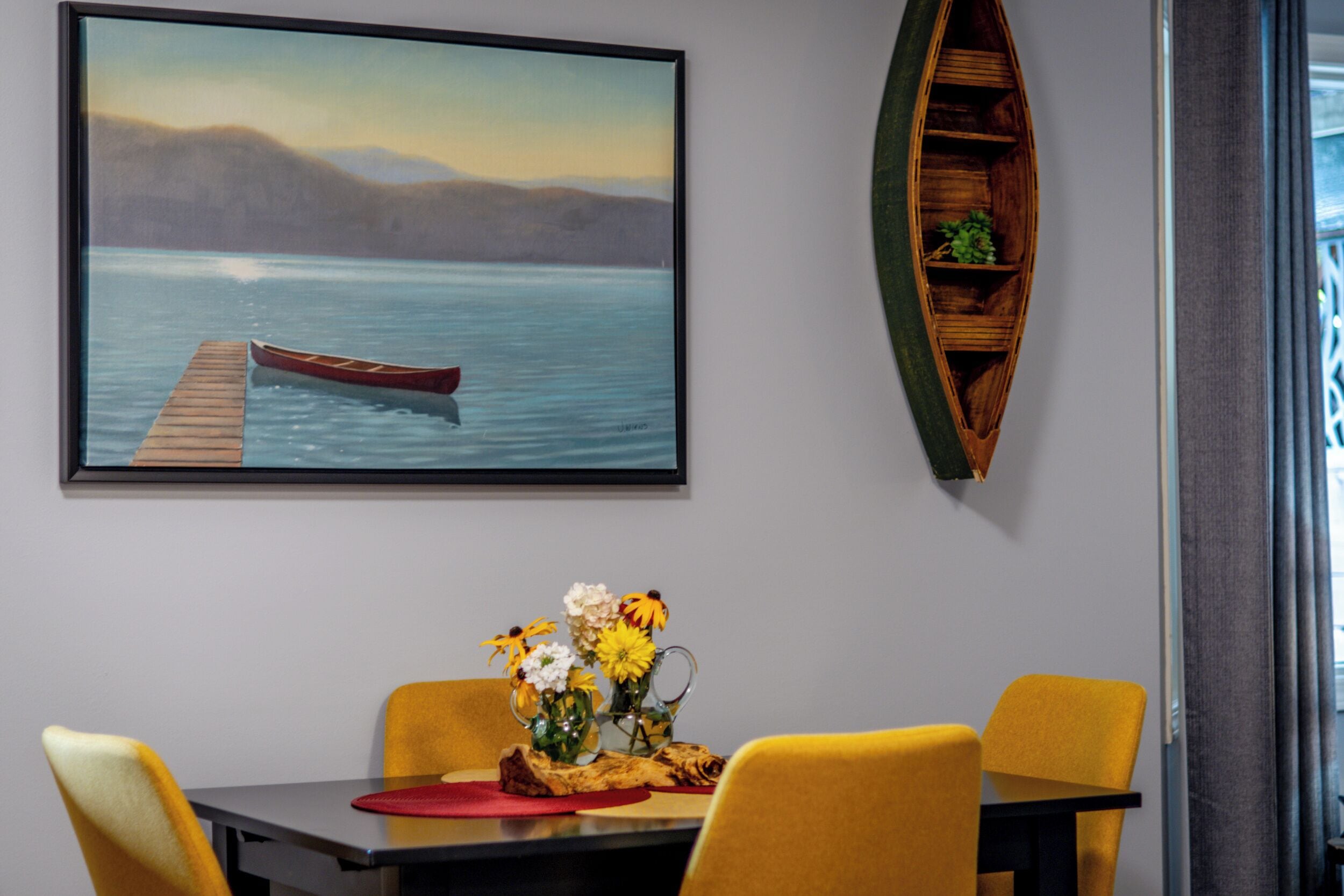 A dining area with a table, chairs, a painting of a lake, and a wooden shelf with a plant.