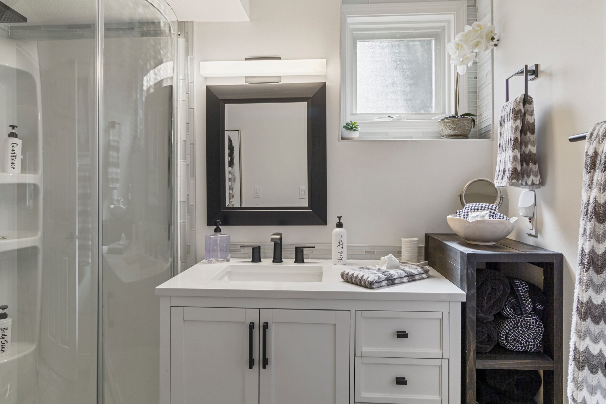 Un baño moderno con cabina de ducha de vidrio, tocador blanco con un espejo grande y una ventana sobre el lavabo.