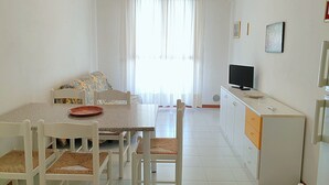 A compact living space with a dining table, chairs, a sofa, a television, and a kitchenette. - Enjoy Your Stay in our Cozy Bright Flat (San Michele al Tagliamento)