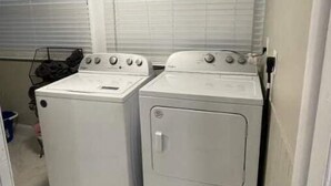 A laundry room with two white washing machines and a dryer, a tiled floor, and a window with blinds. - Comfortable new 2- bedroom house (Jacksonville)