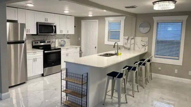 A modern kitchen with white cabinets, a stainless steel refrigerator, a microwave, and a sink. There are bar stools and a small island with a shelf. - Comfortable new 2- bedroom house (Jacksonville)