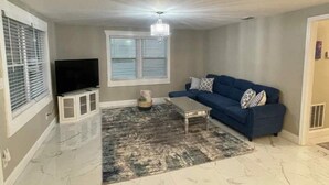 A living room with a blue sofa, a television, a chandelier, and a patterned rug. - Comfortable new 2- bedroom house (Jacksonville)