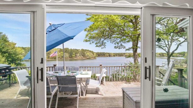 A covered outdoor deck with a table and chairs, overlooking a lake with trees and an American flag. - Waterfront, Incredible Views and Private Dock! (Yarmouth)