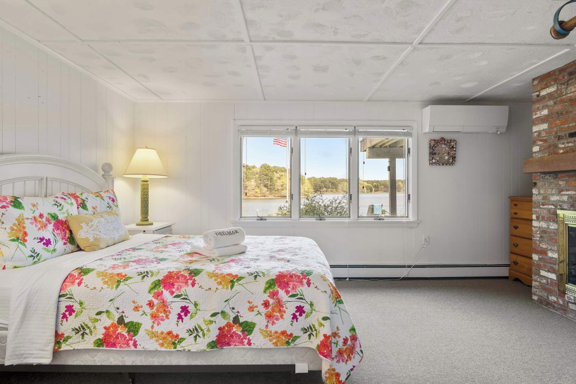 A bedroom with a bed, a nightstand, a lamp, a window with a view of trees and a body of water, and a brick wall.