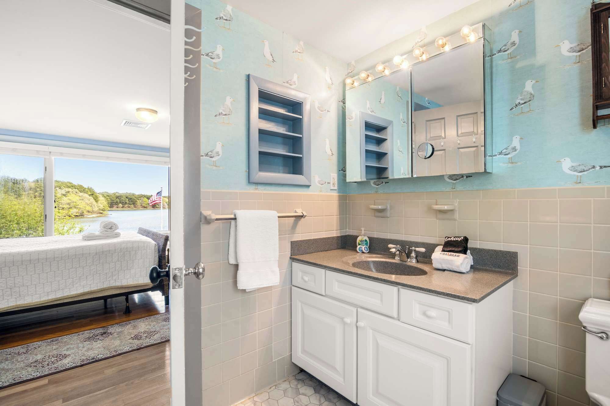 A bathroom with a large mirror, white cabinets, and a view of a lake through an open door.