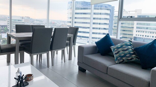A modern living room with a grey sofa, blue and patterned pillows, a dining area with grey chairs and a table, and large windows offering a city view.