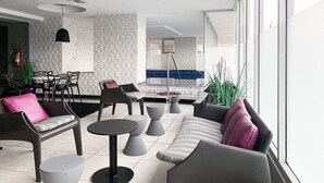 A modern lounge area with sofas, chairs, and tables. There is a large window providing natural light and a view of the outside.