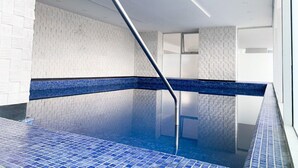 An indoor swimming pool with a blue tiled floor and walls, a metal handrail, and a white brick wall.