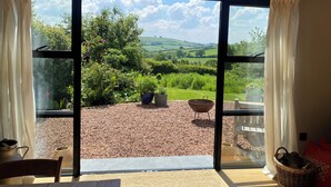 Une pièce dotée d’une grande porte-fenêtre coulissante qui donne sur un espace extérieur comprenant une terrasse en gravier, des plantes en pot et une vue sur des collines verdoyantes.
