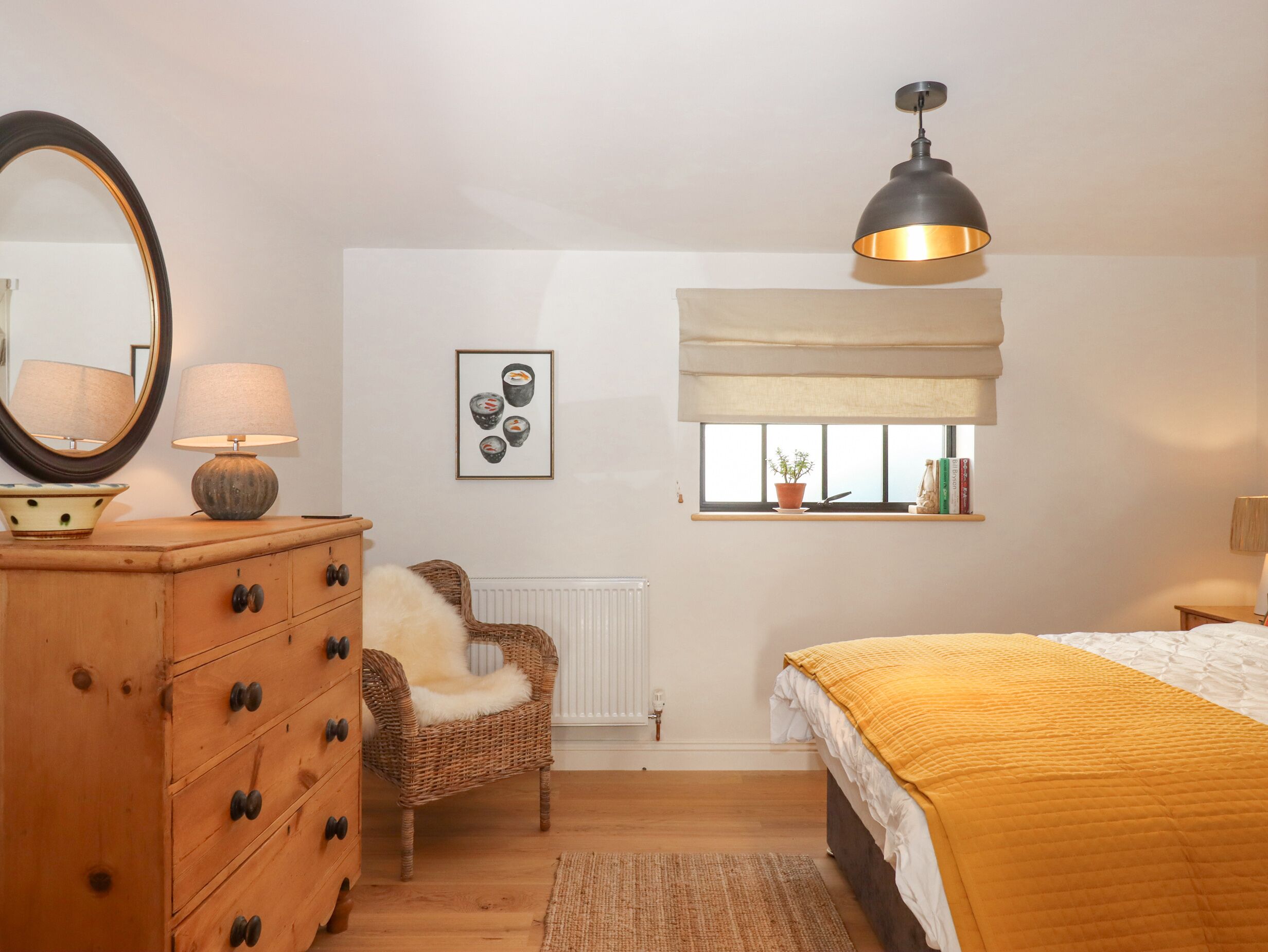 Ein Schlafzimmer mit einem hölzernen Schminktisch, einem Sessel, einem Bett mit gelber Decke, einer Pendelleuchte, einem Fenster mit Pflanze und einem gerahmten Bild an der Wand.