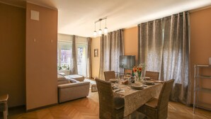 A dining area with a table set for four, surrounded by wicker chairs, a television, and a view of the outside through large curtains. - 3 bedroom nice apartment in Rovereto (Rovereto)