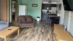 Ein gemütlicher Wohnraum mit Sofa, Couchtisch aus Holz, gemustertem Sessel, Esstisch, Küchenzeile mit Mikrowelle und Deckenventilator.