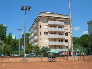 Een meerlaags gebouw met balkons, een tennisbaan en vlaggen.