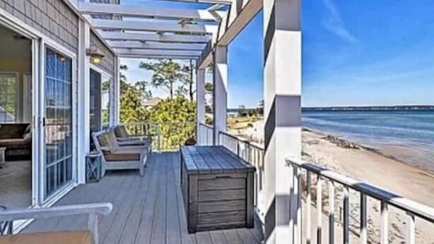Eine Holzdielen-Terrasse mit Glastür zu einem Zimmer, einer Bank und Blick auf den Strand.