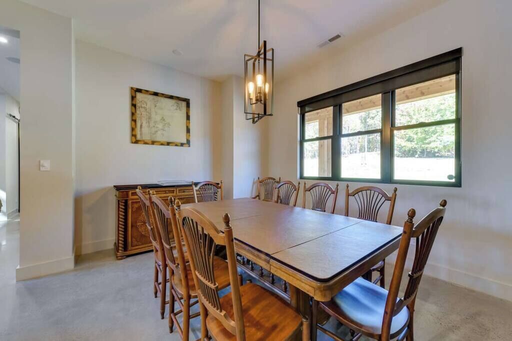 A dining room with a rectangular table and six wooden chairs, a chandelier, a window with a view of greenery, and a framed artwork on the wall.