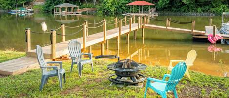 A lakeside deck with chairs, a fire pit, and a dock extending into the water.