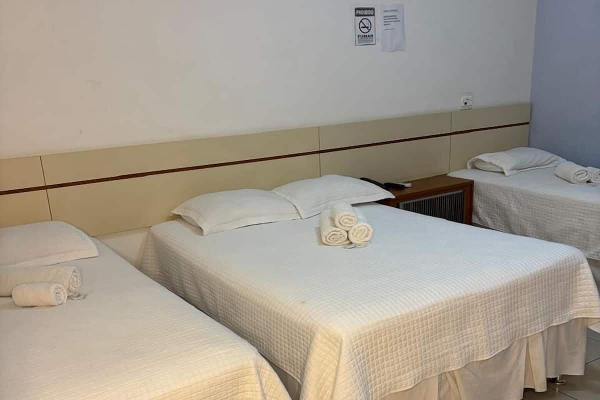 A hotel room with three single beds, each with a white bedspread and pillows. There is a wooden nightstand and a small table with a lamp.