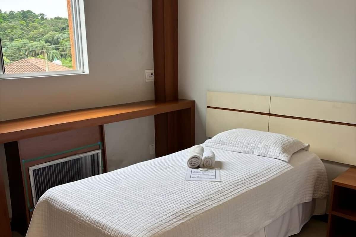 A single bed with a white quilted bedspread and two rolled towels, a wooden headboard, a small wooden nightstand, and a window with a view of greenery outside.
