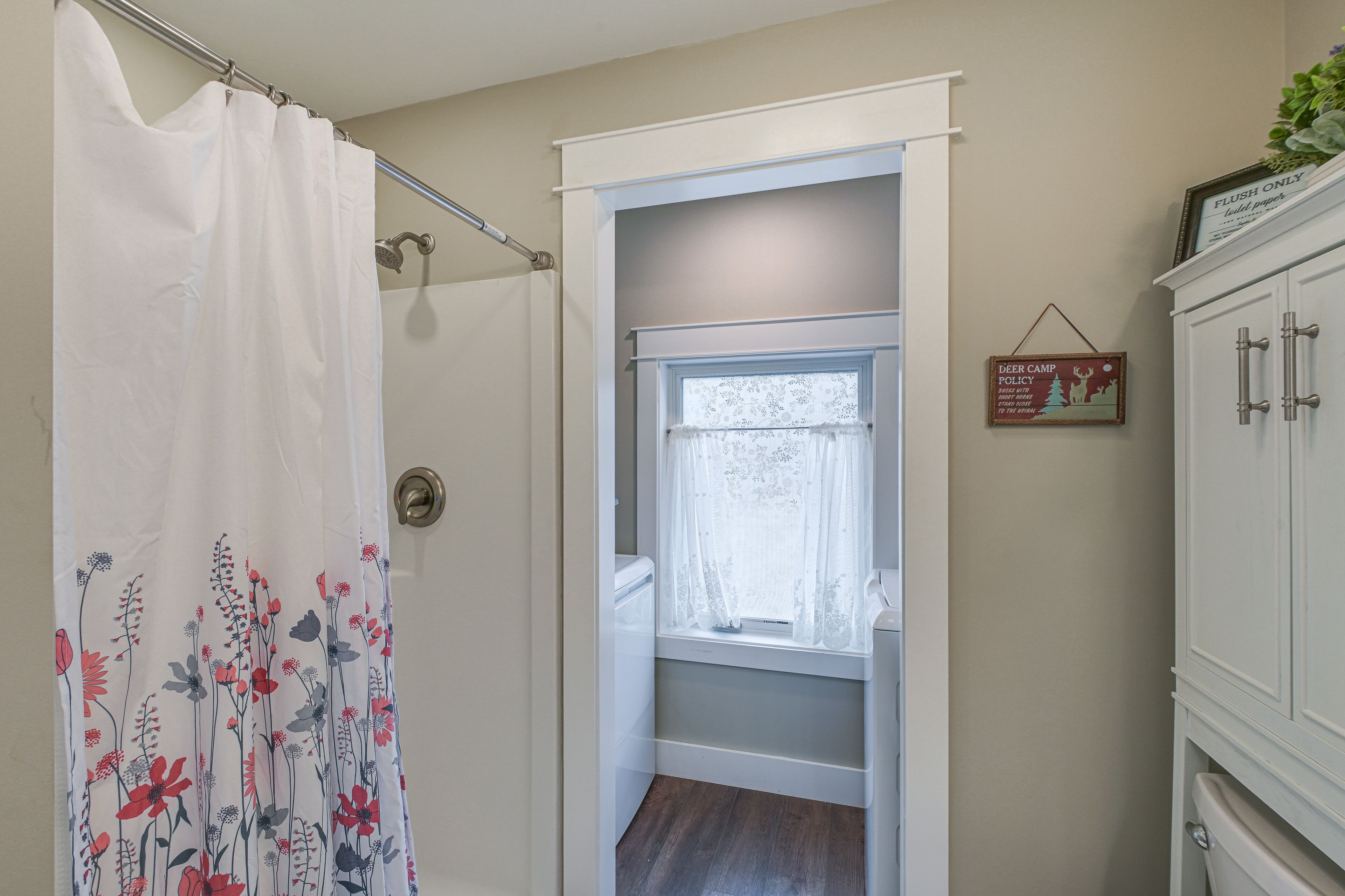 Une salle de bain avec un rideau de douche, une fenêtre ornée de rideaux en dentelle, un plancher en bois et un meuble sur lequel est apposée une pancarte.