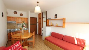 A compact kitchen with wooden cabinets, a dining table with chairs, a red sofa, and a hanging light fixture. - Luminous Apartment on the Shores of Bibione (San Michele al Tagliamento)
