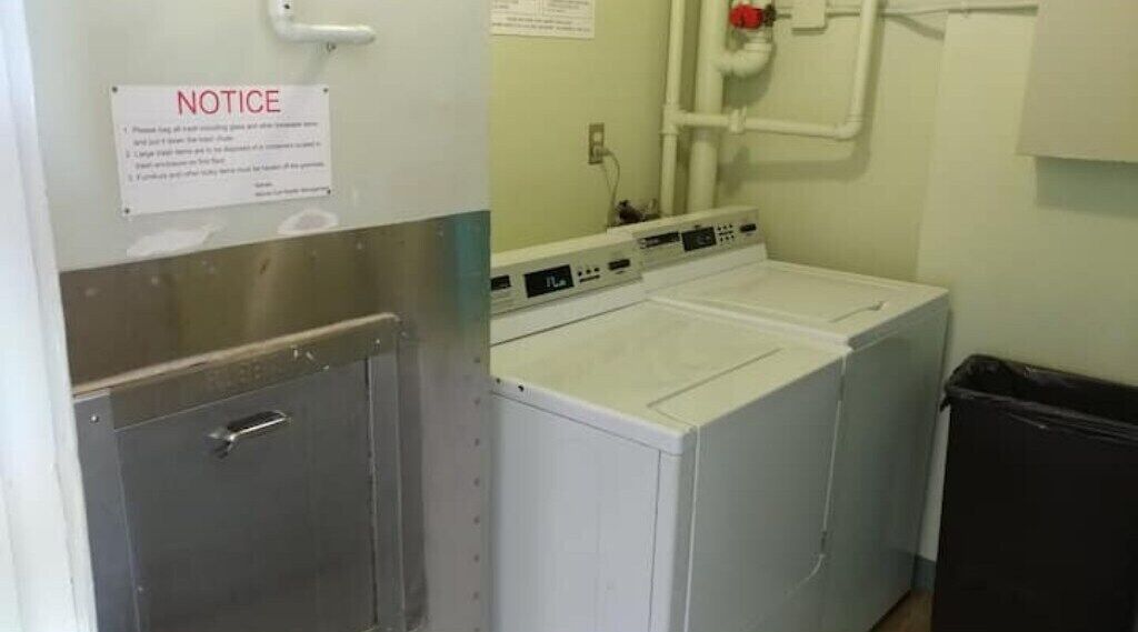 A laundry room with two washing machines, a notice sign, and a trash bin.