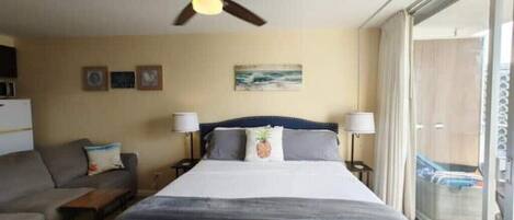A hotel room with a bed, a ceiling fan, a sofa, and a window with a view of the outside.
