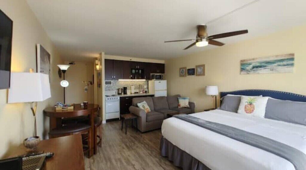 A hotel room with a bed, a desk, a chair, a sofa, a kitchenette, and a ceiling fan.
