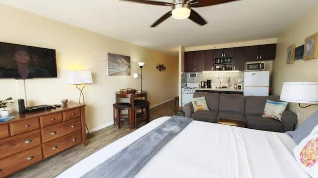 Quarto de hotel compacto com cama, escrivaninha, cadeira, sofá, ventoinha no teto e cozinha compacta ao fundo.