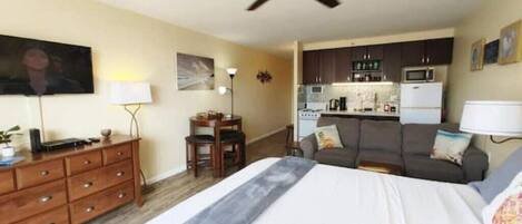 A compact hotel room with a bed, a desk, a chair, a sofa, a ceiling fan, and a kitchenette in the background.