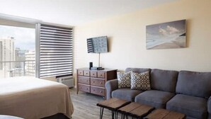 Moderni hotellihuone, jossa on suuri sänky, harmaa sohva, puinen sohvapöytä, jalustalla oleva televisio ja ikkunasta avautuva näkymä kaupunkiin.