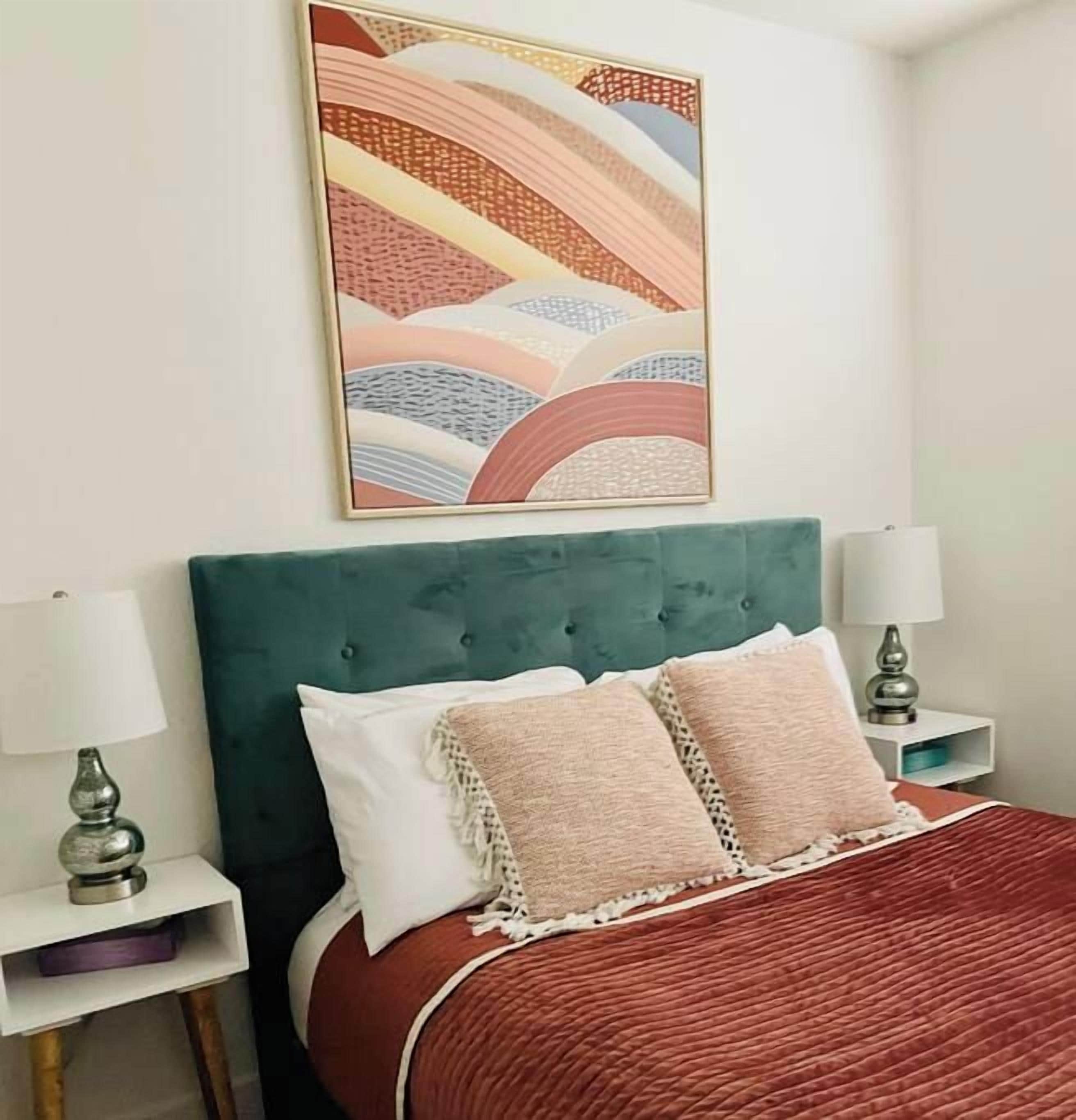 A bed with a green upholstered headboard, two bedside tables with lamps, and a colorful abstract painting above the bed.